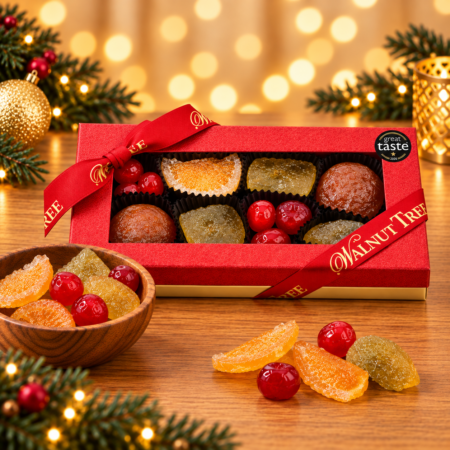 Walnut Tree luxury medium glacé fruit gift box featuring assorted candied fruits, presented in red packaging with a gold-lettered Walnut Tree ribbon, wooden bowl and festive golden Christmas lights.